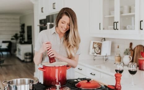 woman holding pepper grinder above a red pot in a kitchen